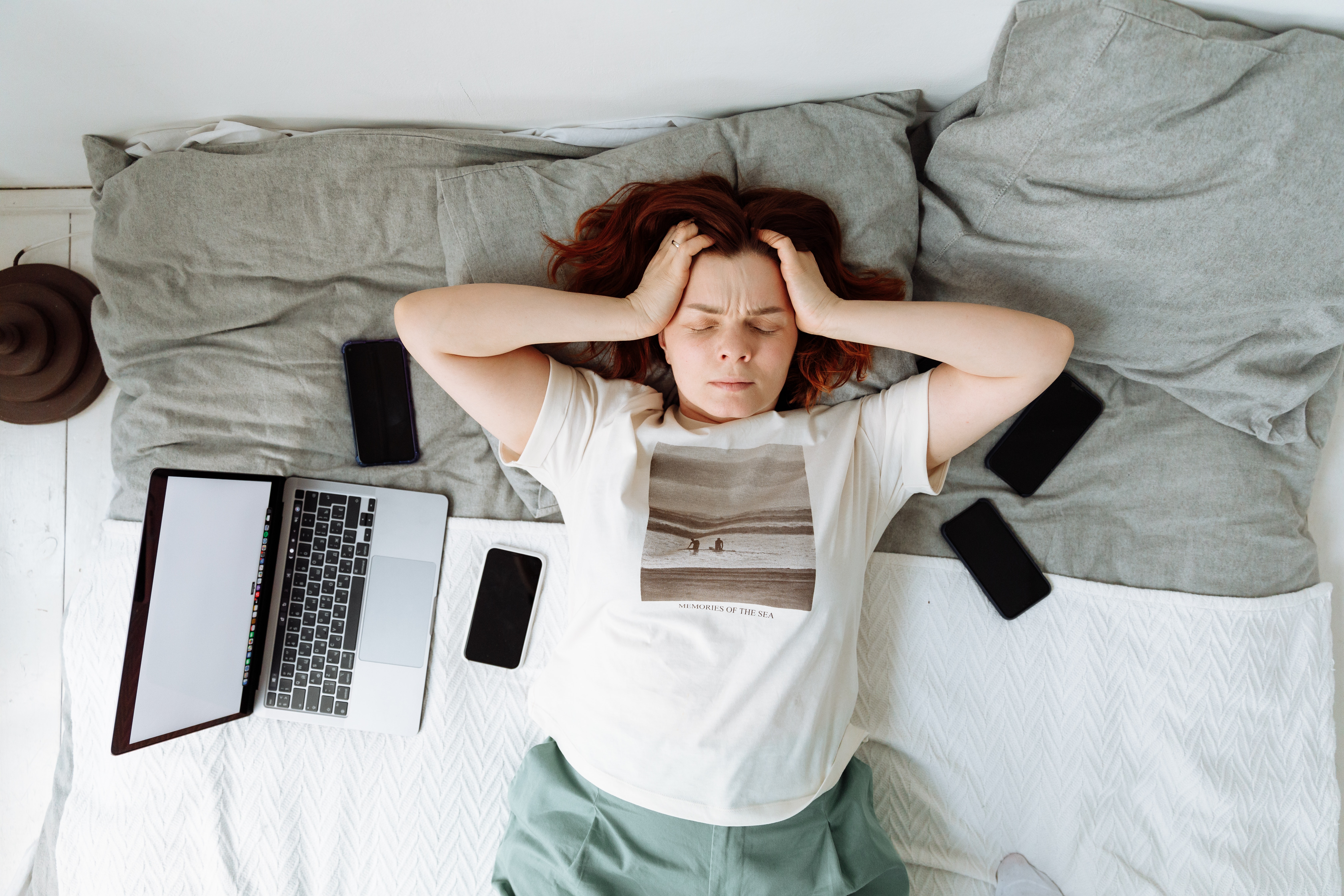 A Stressed Woman Lying on a Bed beside Cellphones and a Laptop